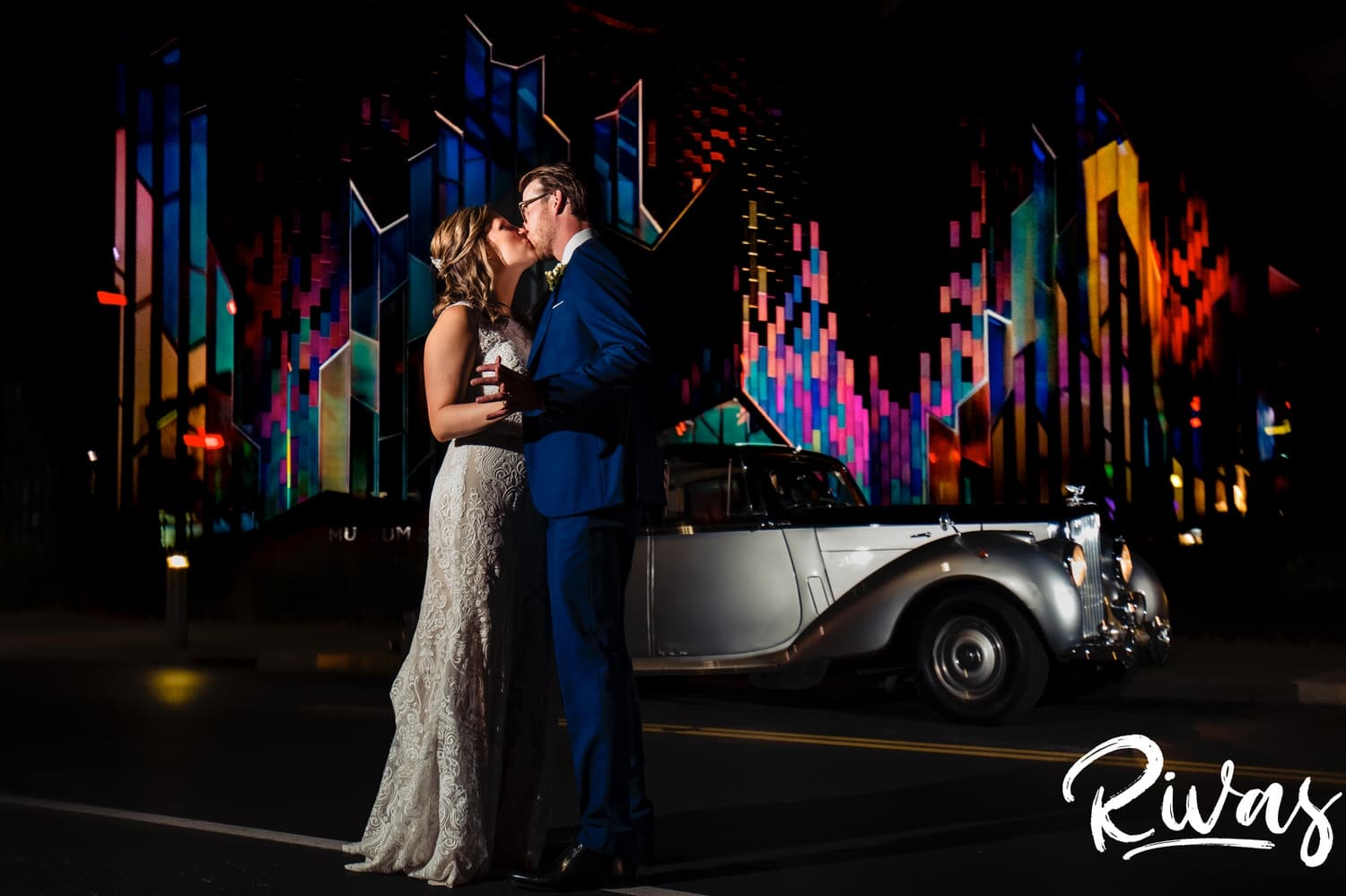 A vibrant picture of a bride and groom holding hands and looking at each other as they stand in front of an antique Bentley parked in front of the Museum at Prairiefire on their wedding night in Kansas City. 