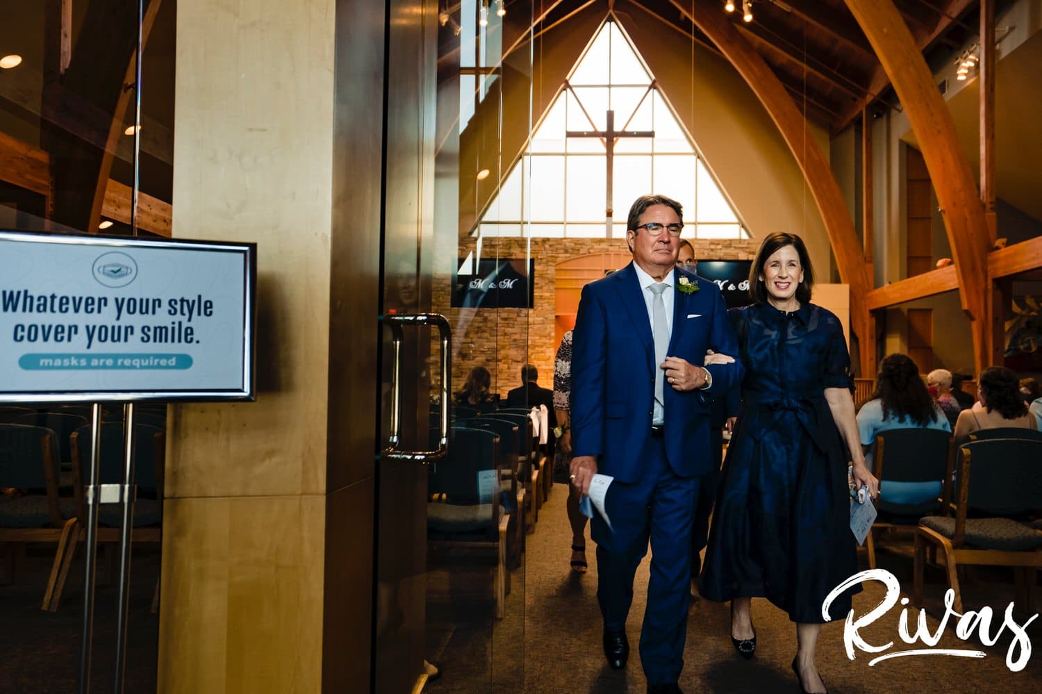 A candid picture of a groom's mom and dad exiting a wedding ceremony with a "wear a mask" sign right by the door. 