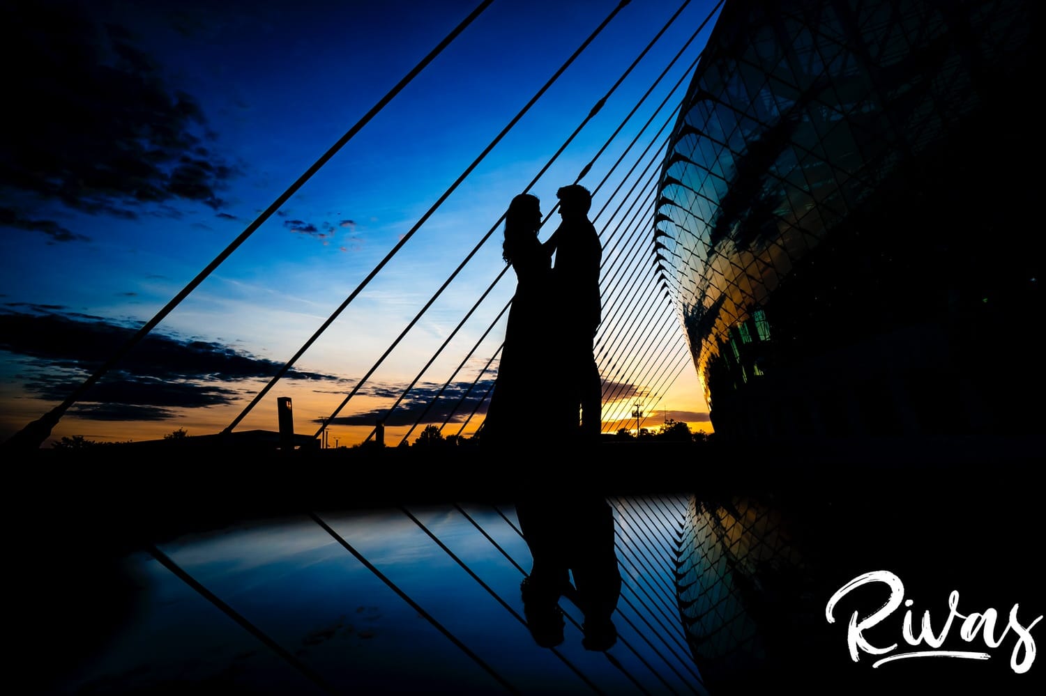 A vibrant, silhouette of an engaged couple sharing an embrace at sunset outside the Kauffman Center in Kansas City, their reflection visible on the ground beneath them. 