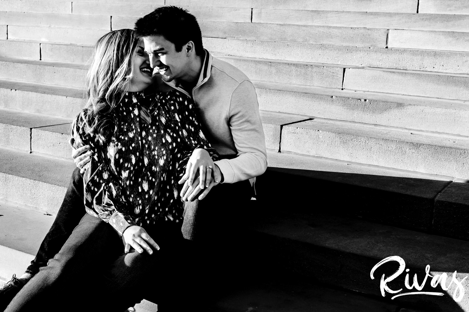 A bold black and white, candid engagement picture of an engaged couple sitting on a set of concrete stairs, laughing and whispering together during their fall engagement session at The Nelson Atkins Museum of Art in Kansas City. 