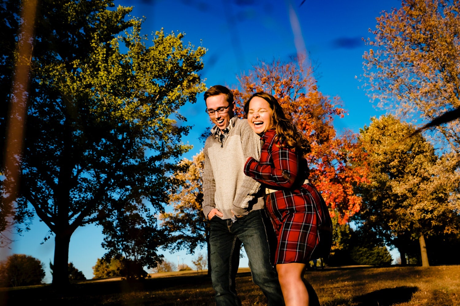 A colorful, vibrant picture of a woman in a red plaid dress tickling a man in a gray sweater against a backdrop of blue skies and colorful trees during their fall engagement session in Kansas City. 