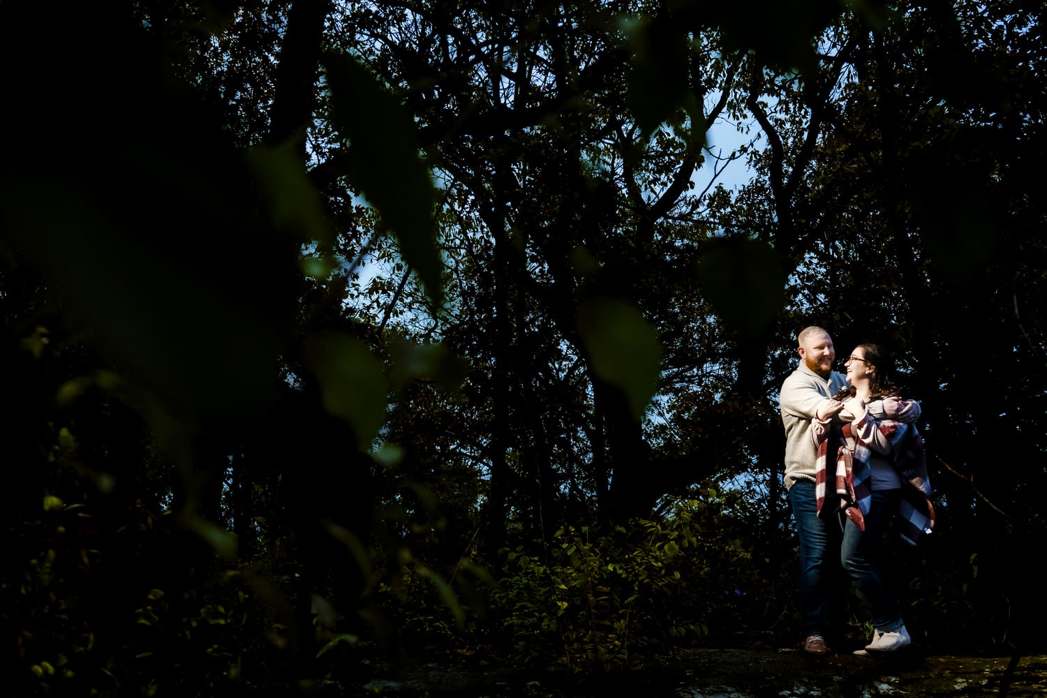 A dark and moody portrait of an engaged couple standing on a cliffside in fall sweaters, surrounded by green leaves and trees during their moody fall engagement session in Kansas City. 