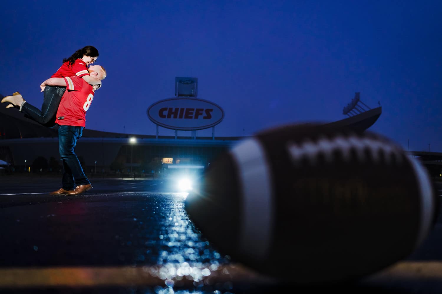 A vibrant, moody picture of an engaged couple in Chief's jerseys sharing an embrace in front of Arrowhead Stadium in the background of an image, with a football visible in the foreground of the image during a moody fall engagement session in Kansas city. 