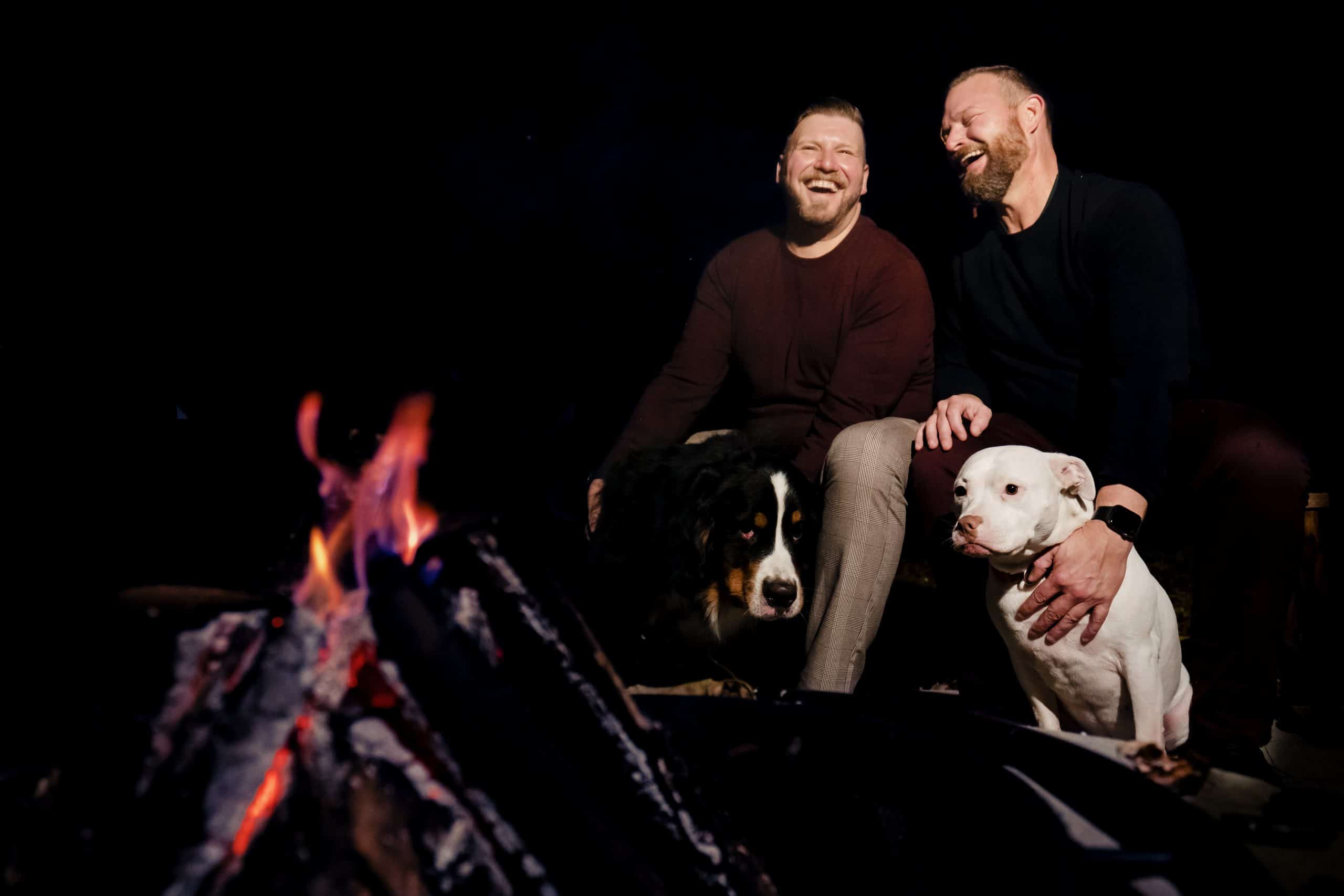 An image taken up-close to a fire, of two men in fall sweaters sitting on a bench, laughing hysterically together as they pet their dogs at the end of their outdoorsy fall engagement session in Kansas City. 