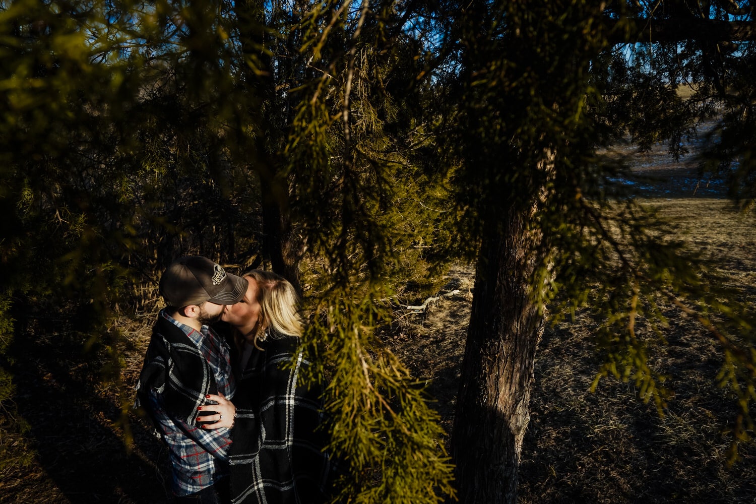 A colorful, candid picture taken through the limbs of a tree of a couple leaning in to share a kiss during a winter engagement session. 