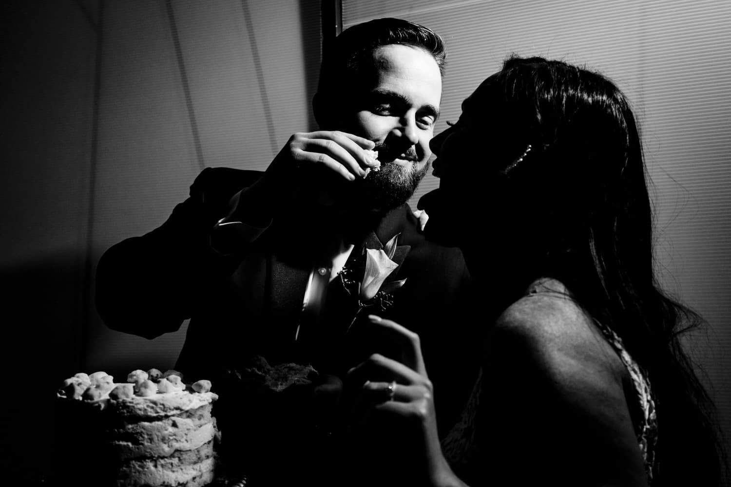 A candid black and white picture of a groom feeding a bride a piece of cake during their 2020 wedding reception in KC. 