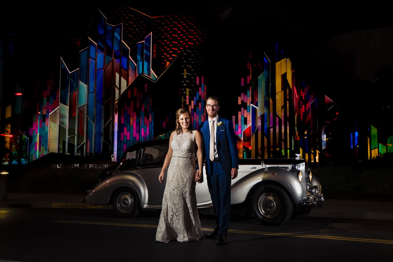 A colorful, candid picture of a bride and groom holding hands and walking away from the Museum at Prairiefire and a classic car on their 2020 wedding day in KC. 