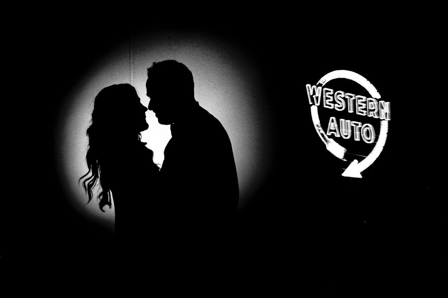A black and white silhouette of a bride and groom leaning in to share a kiss, the Western Auto sign visible in the background at the end of their 2020 KC wedding day at The Otten on Main. 
