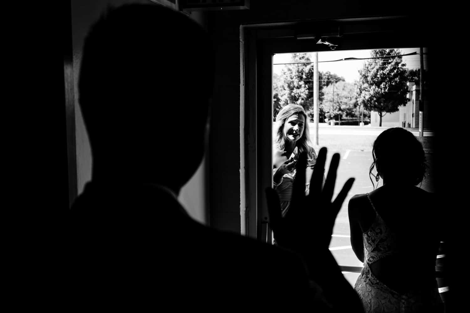 A candid, black and white picture of a bride and groom waving at a bride's sister through a glass door during a 2020 wedding in KC. 