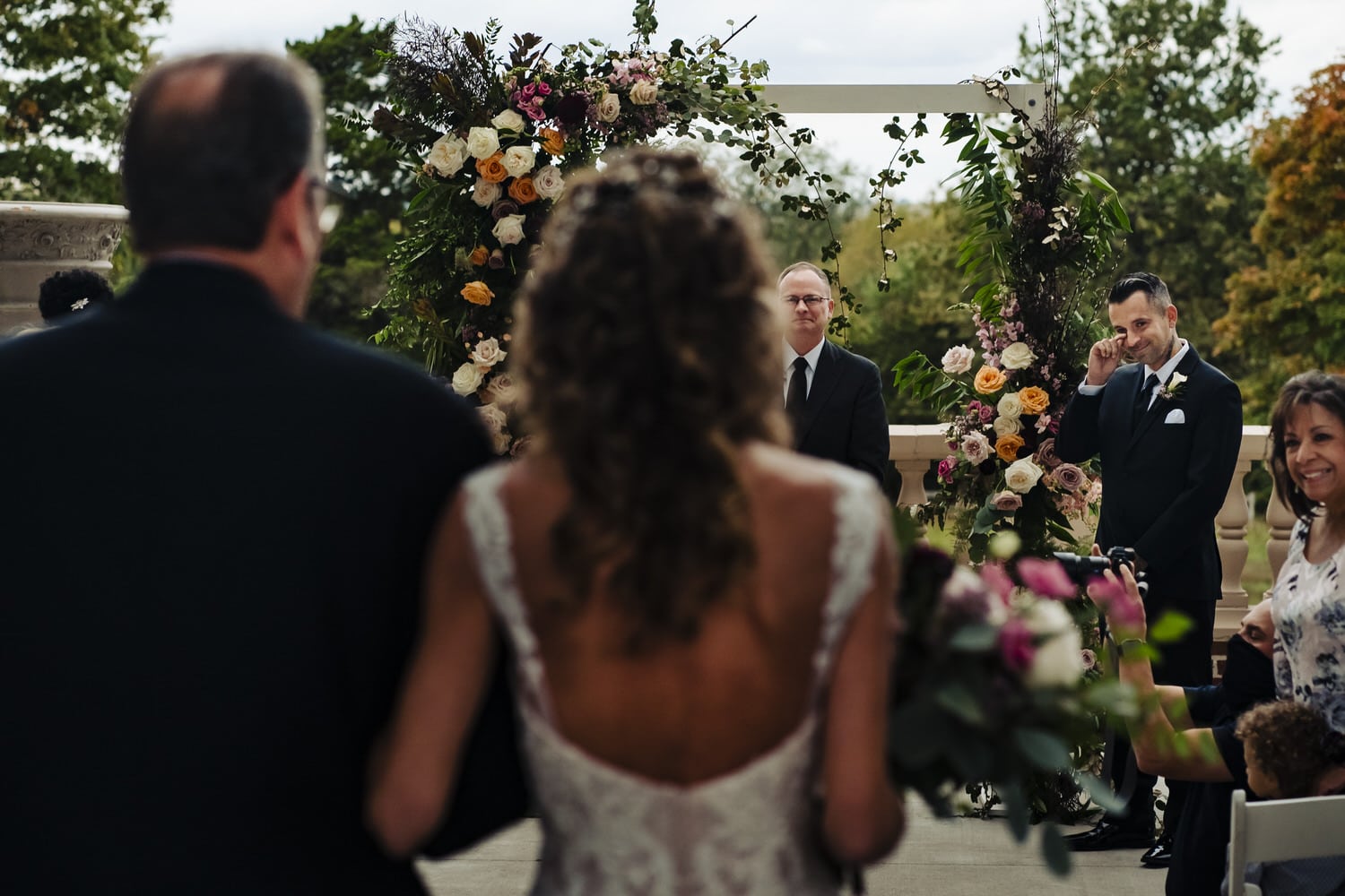 A colorful, candid picture of a bride and her dad walking down the aisle towards her groom, as he wipes tears from his eyes during a 2020 wedding ceremony at Longview Mansion. 