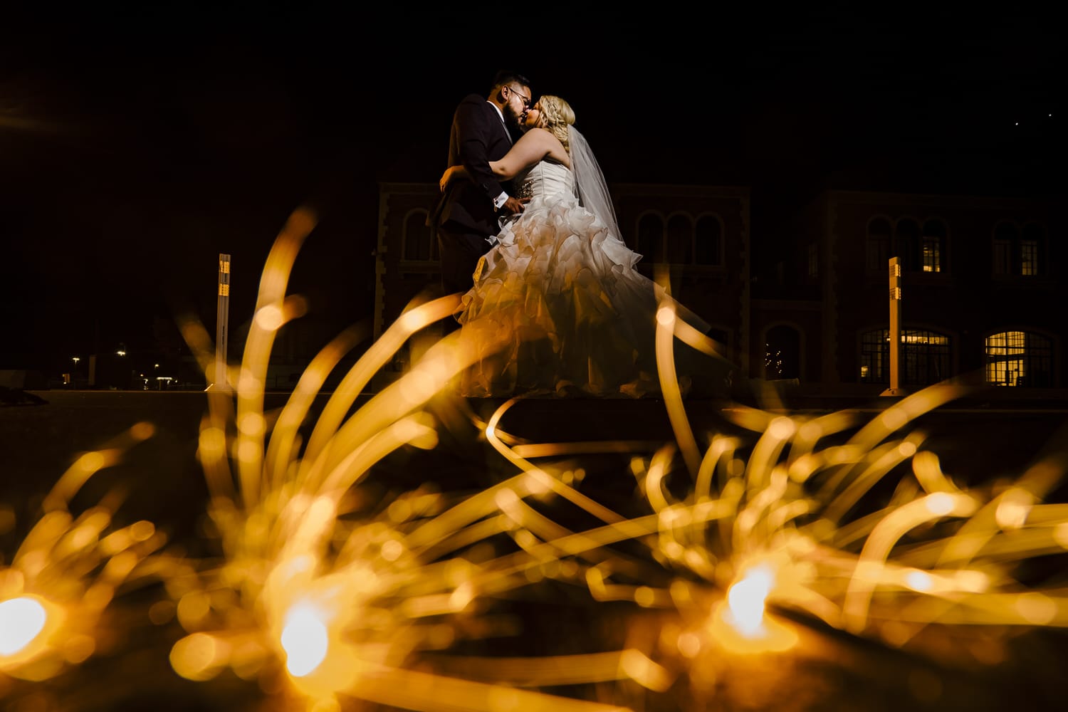 A colorful picture of a bride and groom leaning in to share a kiss as a pile of sparklers burns on the ground in front of them on their wedding day in Kansas City at The DeLeon. 