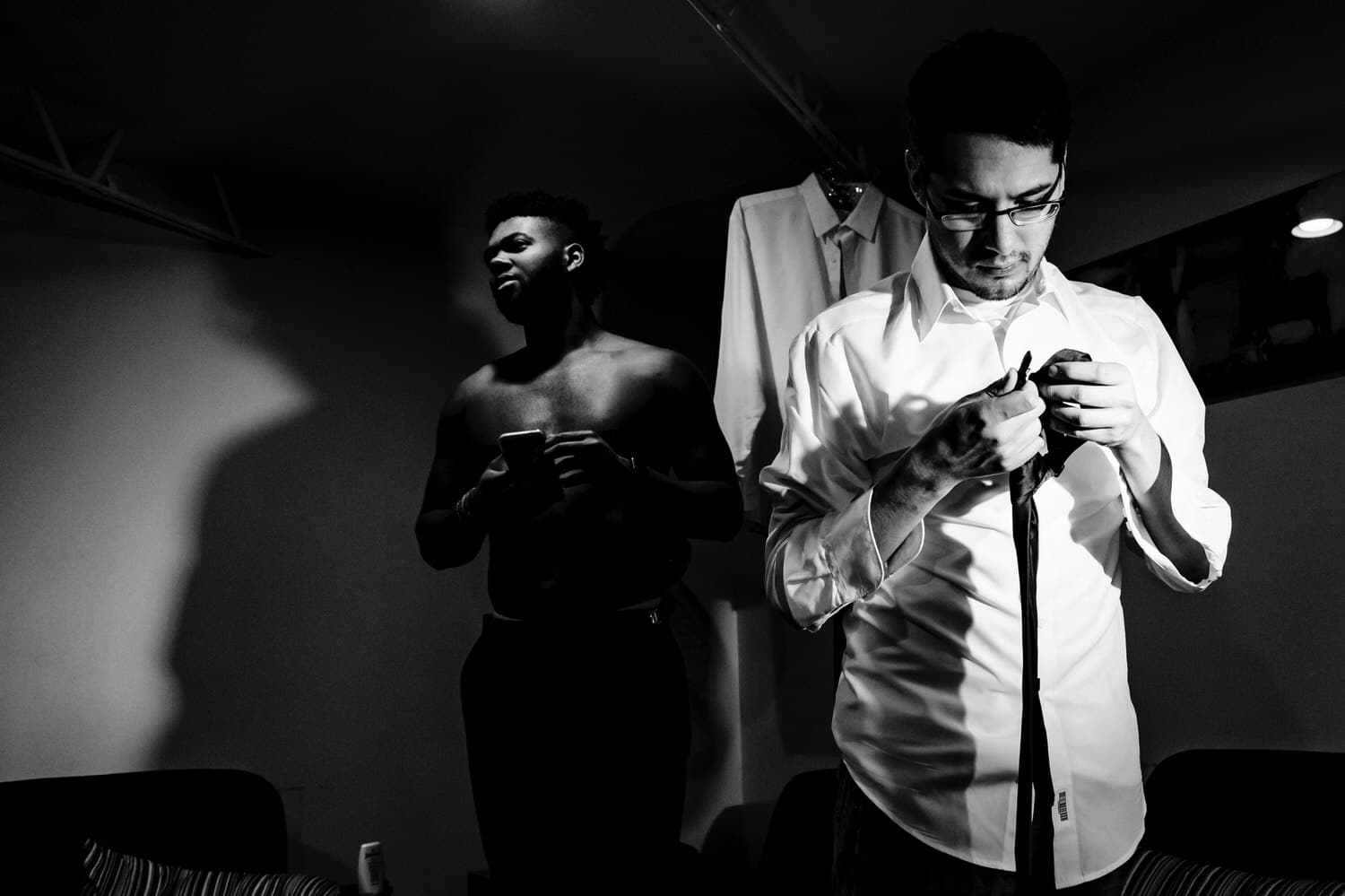 A dramatic black and white picture of two men, one shirtless looking at his cell phone, the other working on tying his tie on the afternoon of a rainy wedding day in Kansas City. 