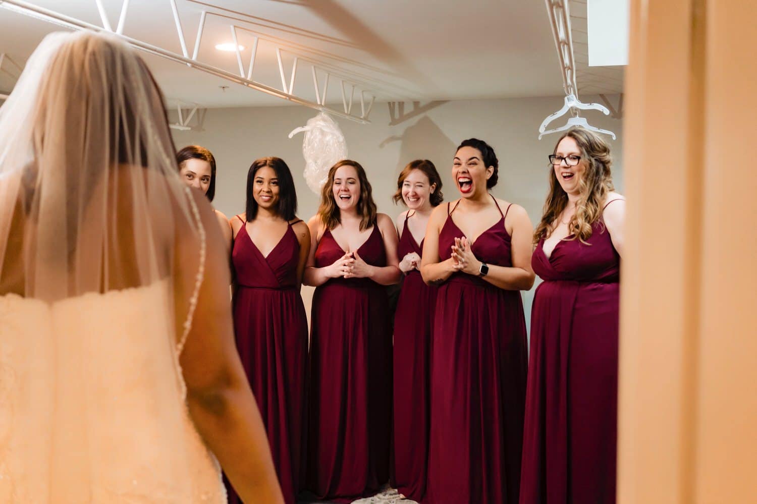 A colorful candid picture of a bride walking out of her dressing room to the look of surprise and joy on her bridesmaids' faces on a rainy winter wedding day at The Station in Kansas City. 