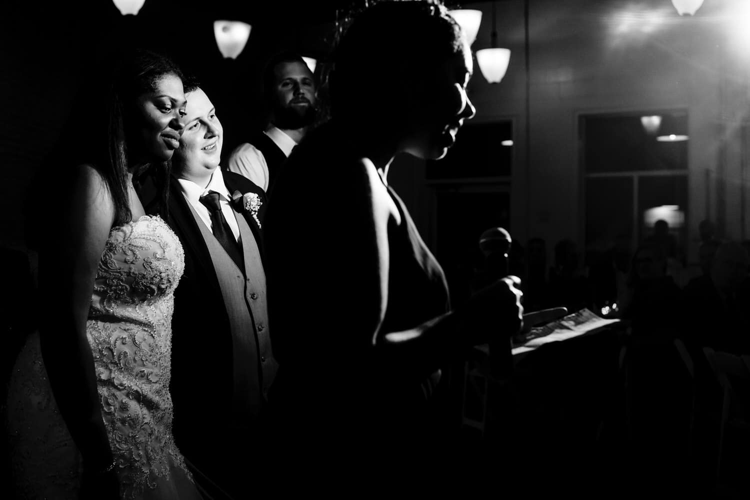 A candid black and white picture of a maid of honor toasting the bride in the foreground, with the image focus on the bride and groom emracing and listening in the background during their wedding reception at The Station in Kansas City.