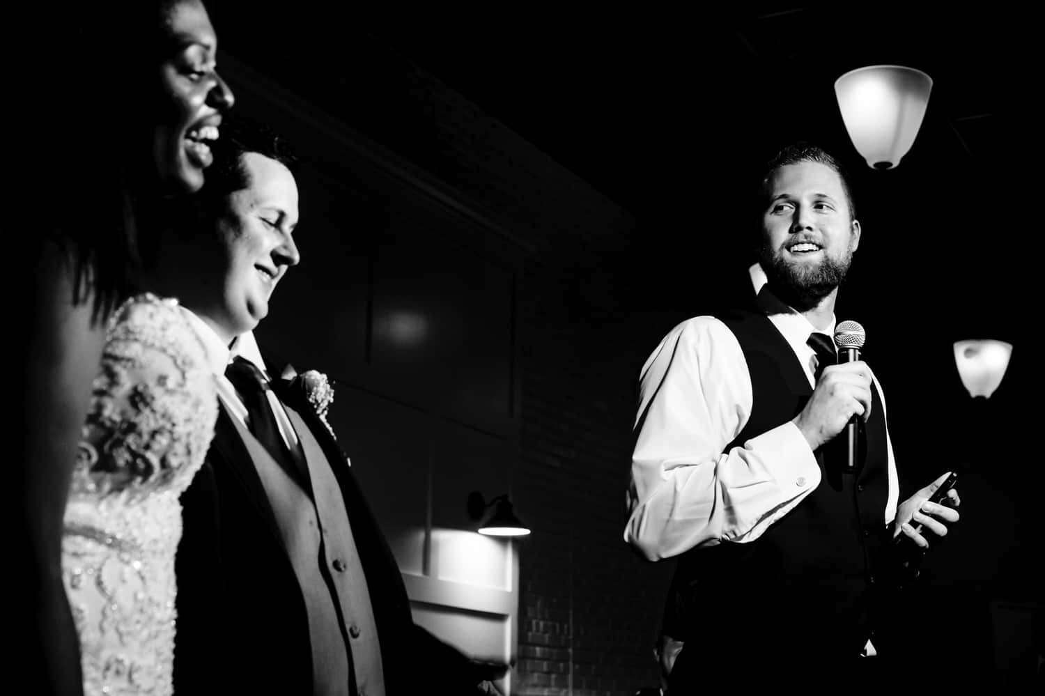 A candid black and white picture of a best man toasting the bride in the background, with the bride and groom embracing and listening in the foreground during their wedding reception at The Station in Kansas City.