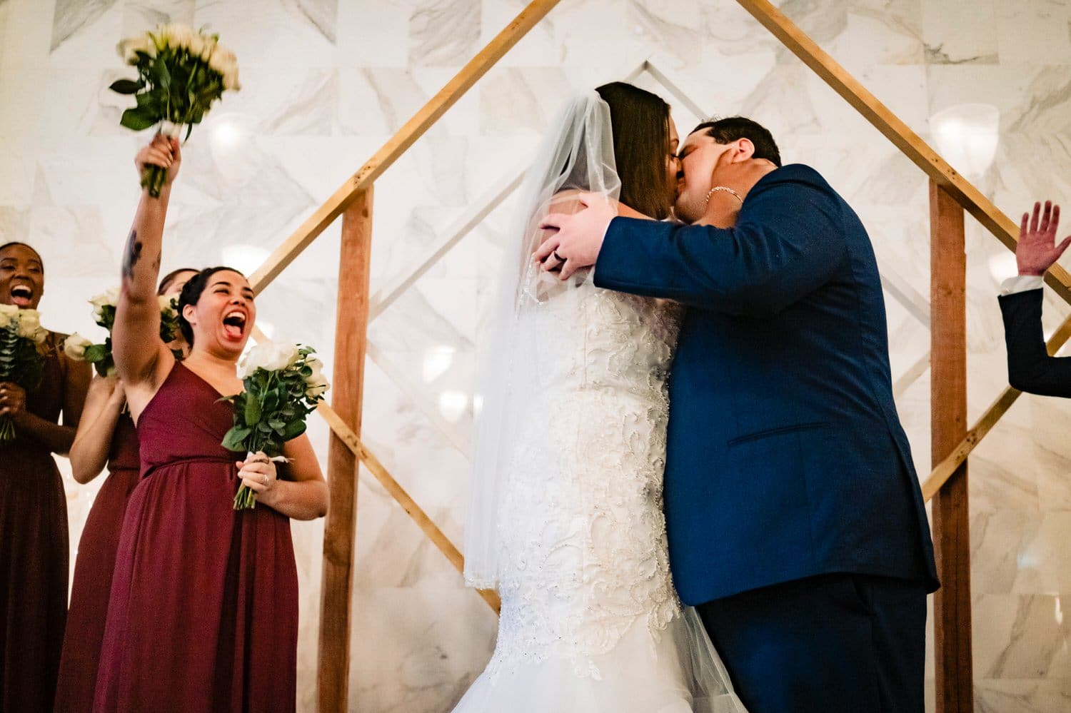 A colorful, candid picture of a bride and groom sharing their first kiss as the maid of honor throws her arms up in celebration at the end of a winter wedding ceremony in Kansas City at The Station. 