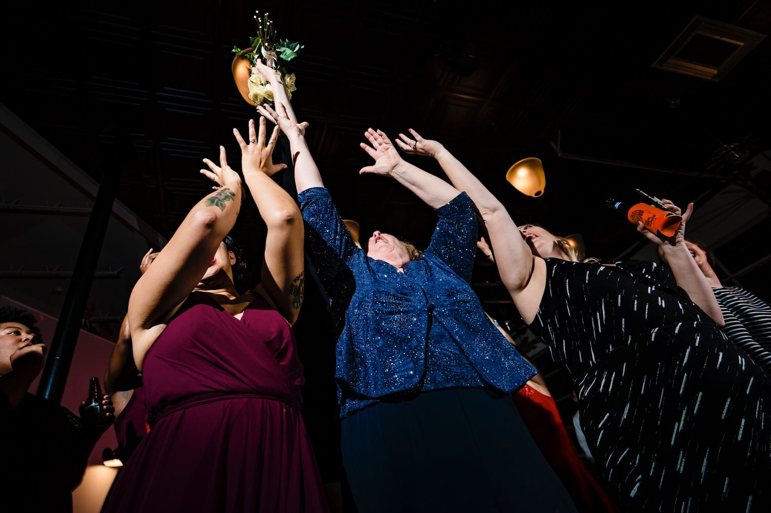 A colorful, candid picture taken from the ground looking up of a group of women reaching up to grab a bouquet that is flying through the air during a winter wedding reception at The Station in Kansas City. 