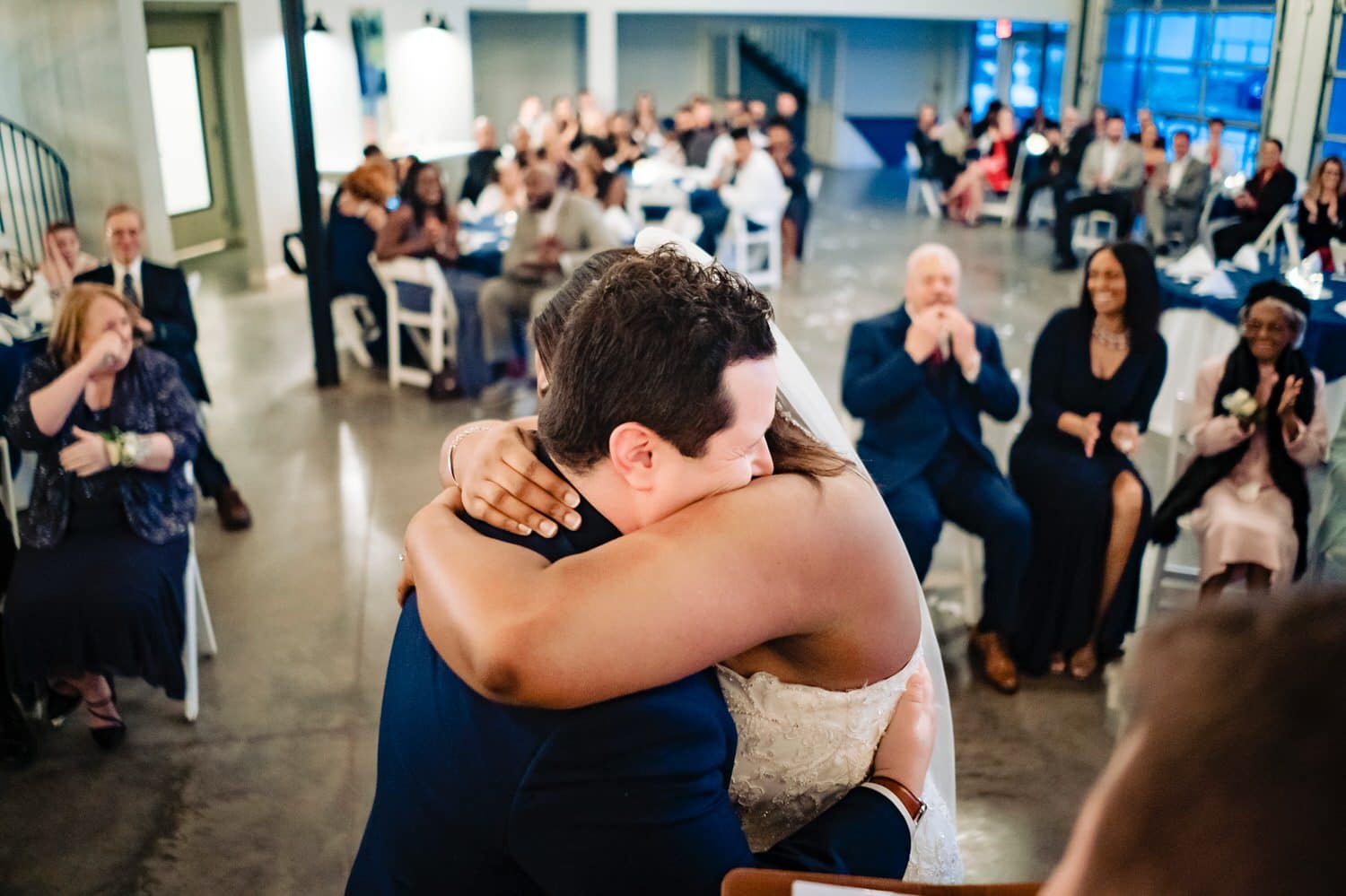 A colorful, candid picture, taken from behind the bride and groom, of them sharing a big hug just after their first kiss, their family and friends cheering them on in the background on a rainy winter wedding day at The Station in Kansas City. 