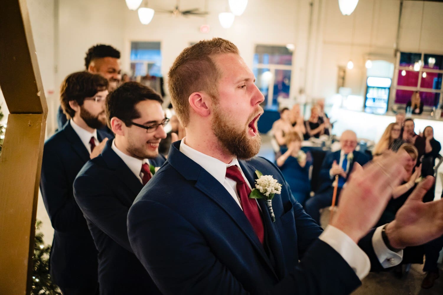 A colorful, candid picture of a best man shouting with joy and clapping enthusiastically at the end of a winter wedding ceremony at The Station in Kansas City. 