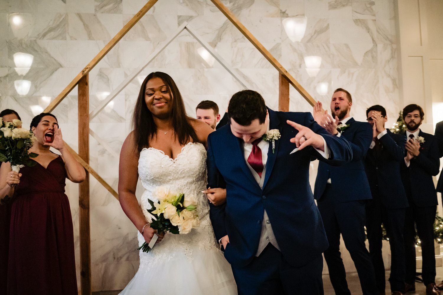 A colorful, candid picture of a bride and groom doing a little bow at the end of their wedding ceremony, as their wedding party clap and shout enthusiastically in the background on their rainy winter wedding day at The Station in Kansas City. 