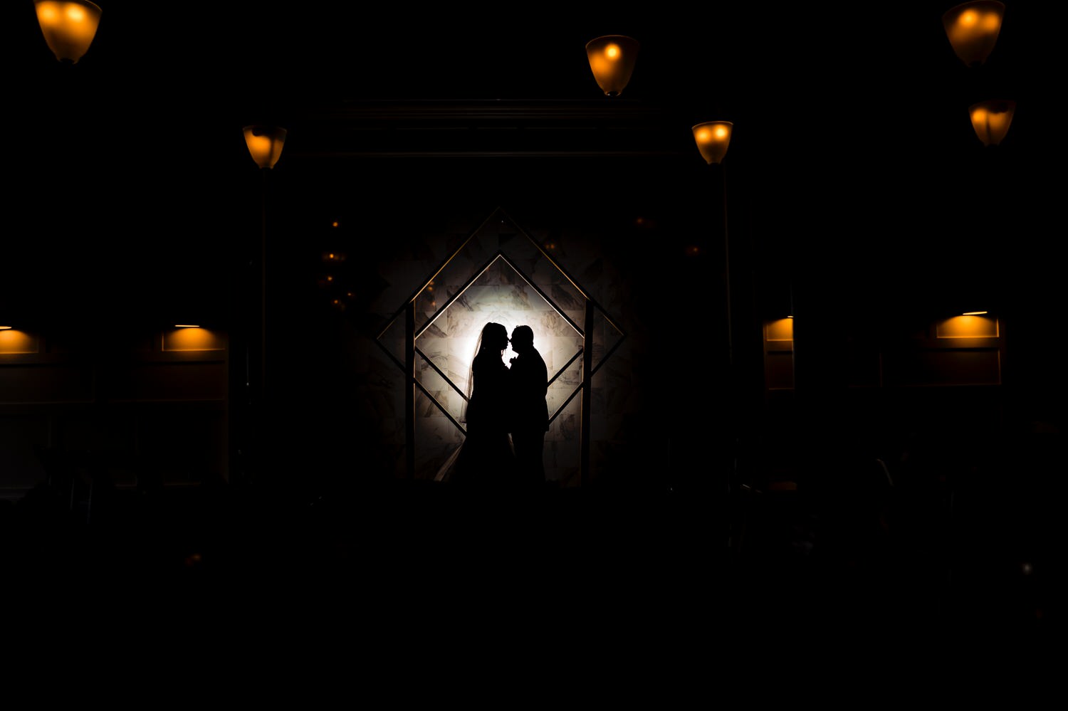A dramatic, colorful silhouette of a bride and groom leaning in to share a kiss as the chandeliers of the venue glow on a rainy winter wedding day in Kansas City at The Station. 