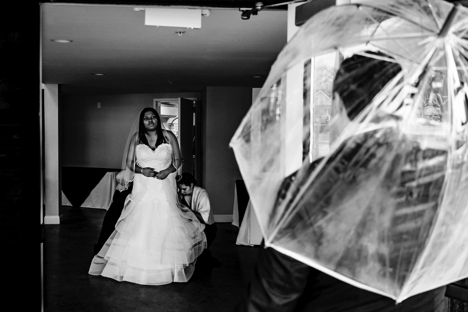 A candid black and white picture of a bride getting her gown bustled as her groom holds an umbrella, watching with interest on a rainy winter wedding day in Kansas City. 