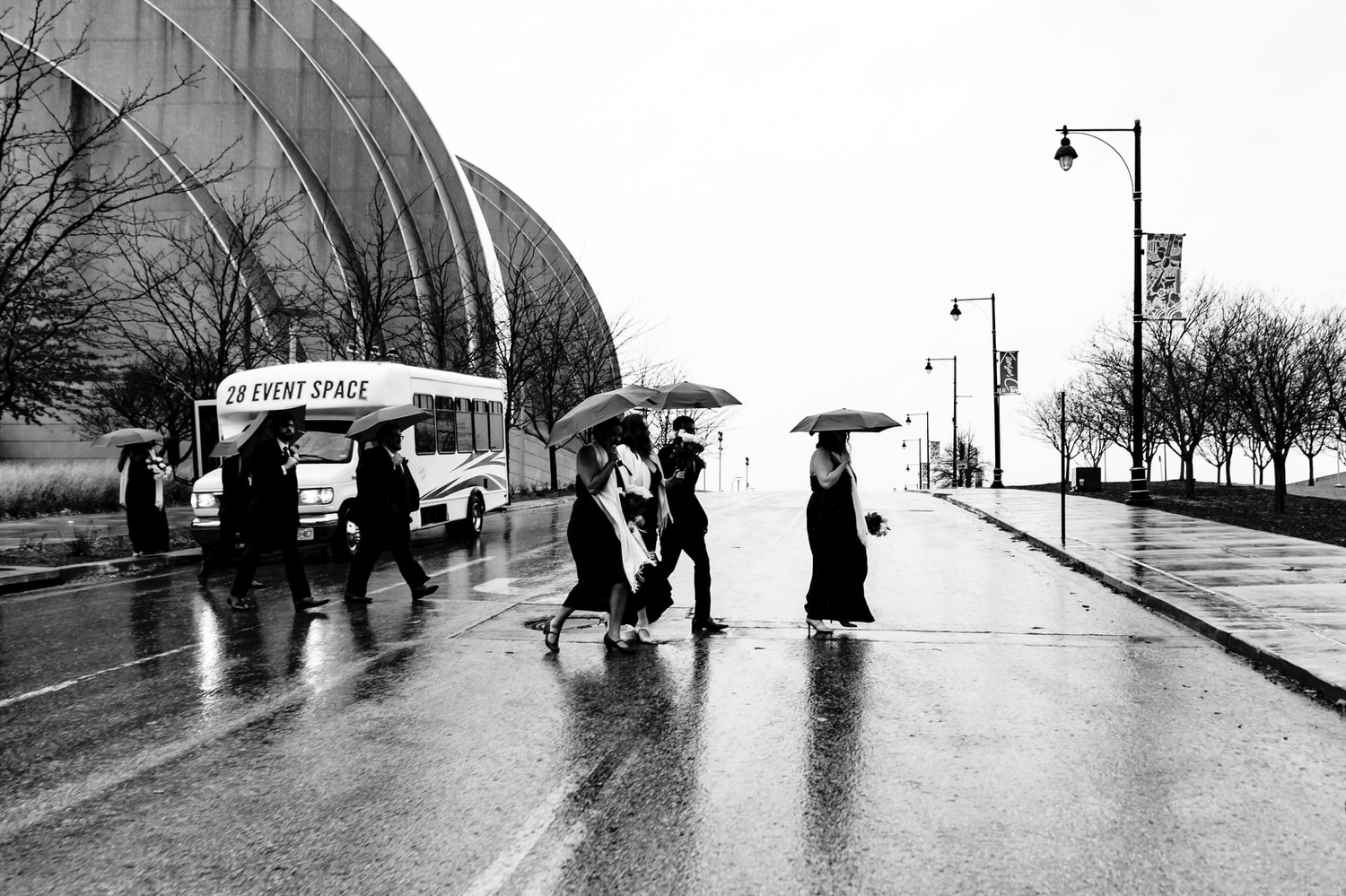 A candid black and white picture of a wedding party holding umbrellas crossing the street in front of the Kauffman Center for the Performing Arts on a rainy winter wedding day in Kansas City. 
