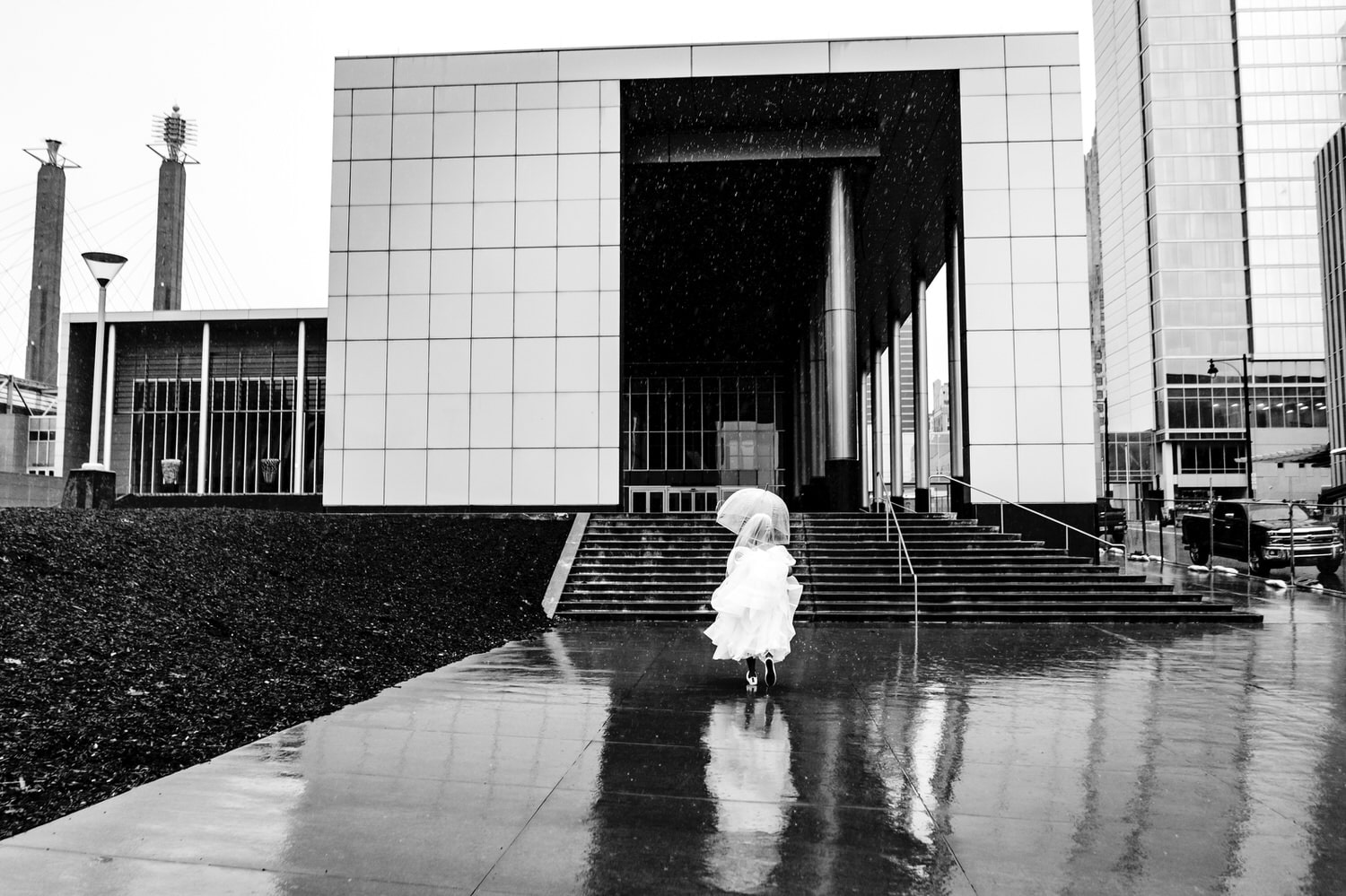 A candid black and white picture of a bride carrying an umbrella, holding her gown up around her knees running up a set of stairs on her rainy wedding day in Kansas City. 