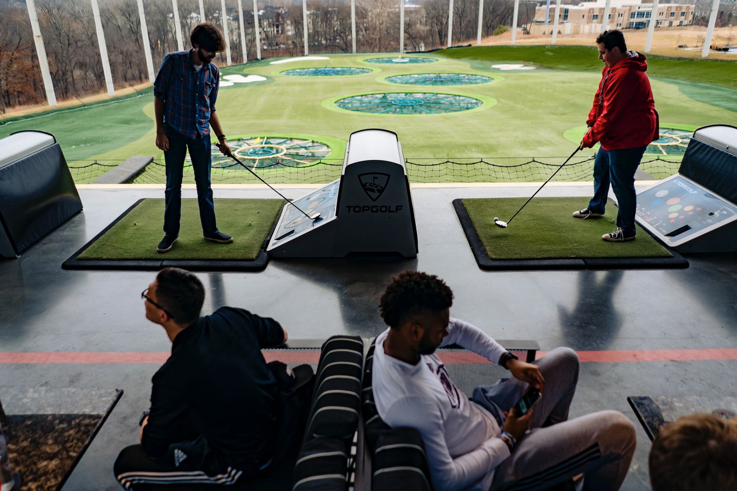 A colorful, candid picture of two men teeing up to hit a golf ball as a group of others watch on, on the morning of a rainy winter wedding at the Kansas City venue, The Station. 