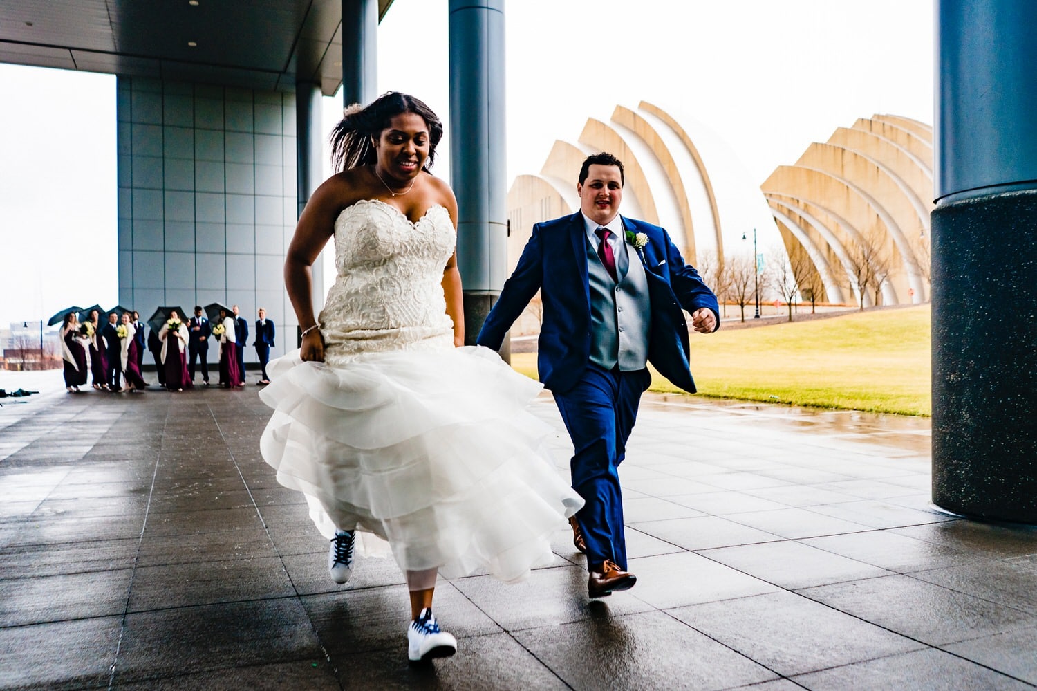 A colorful, candid picture of a bride and groom racing towards the camera, their wedding party visible in the background on a rainy winter wedding day in downtown Kansas City. 