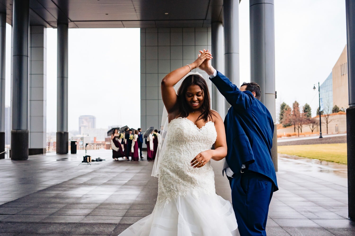A colorful, candid picture of a bride and groom dancing, their wedding party visible in the background on a rainy winter wedding day in downtown Kansas City. 