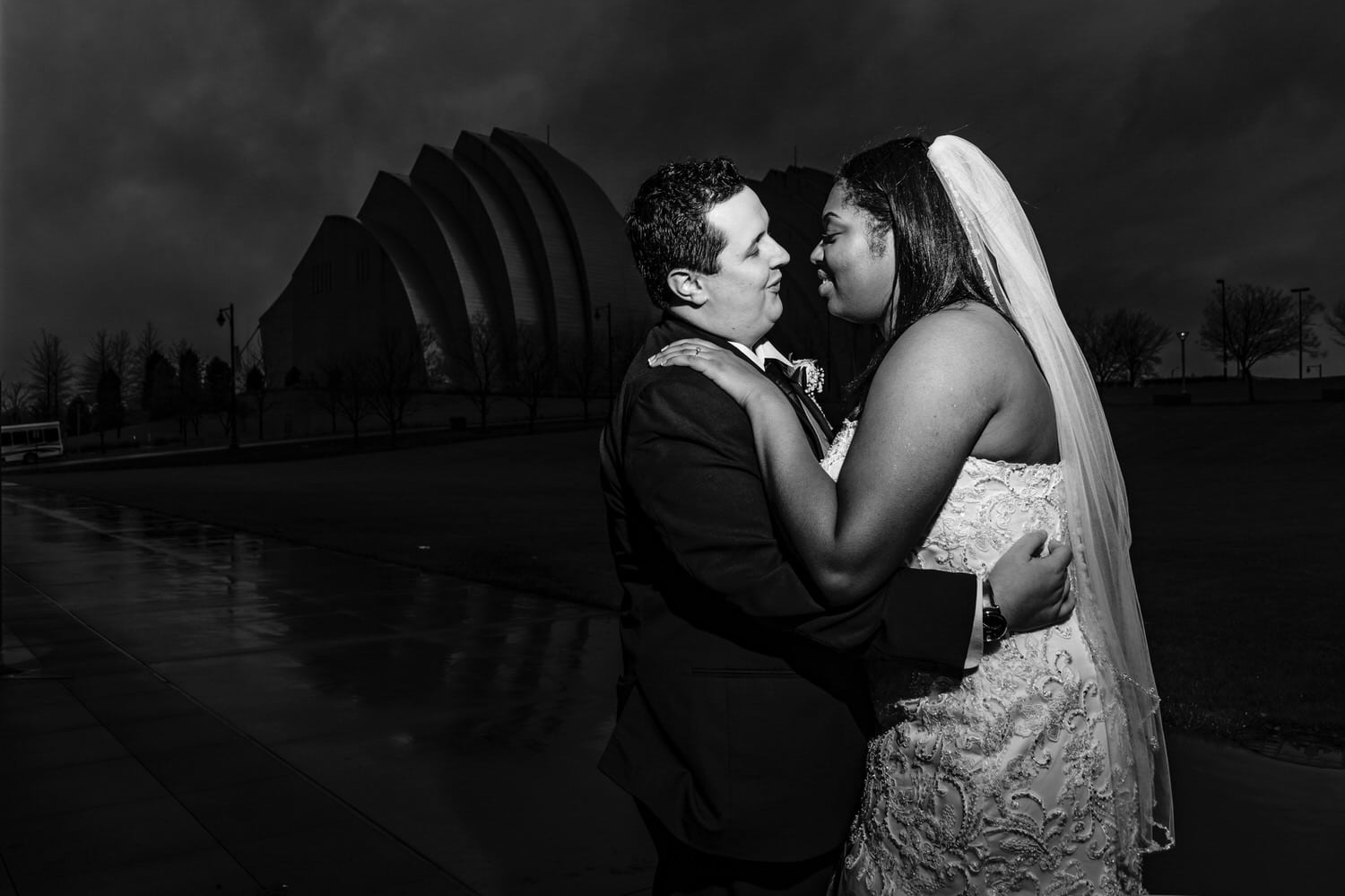 A black and white portrait of a bride and groom standing in front of the Kauffman Center for the Performing Arts leaning in to share a kiss on their rainy winter wedding day in downtown Kansas City. 