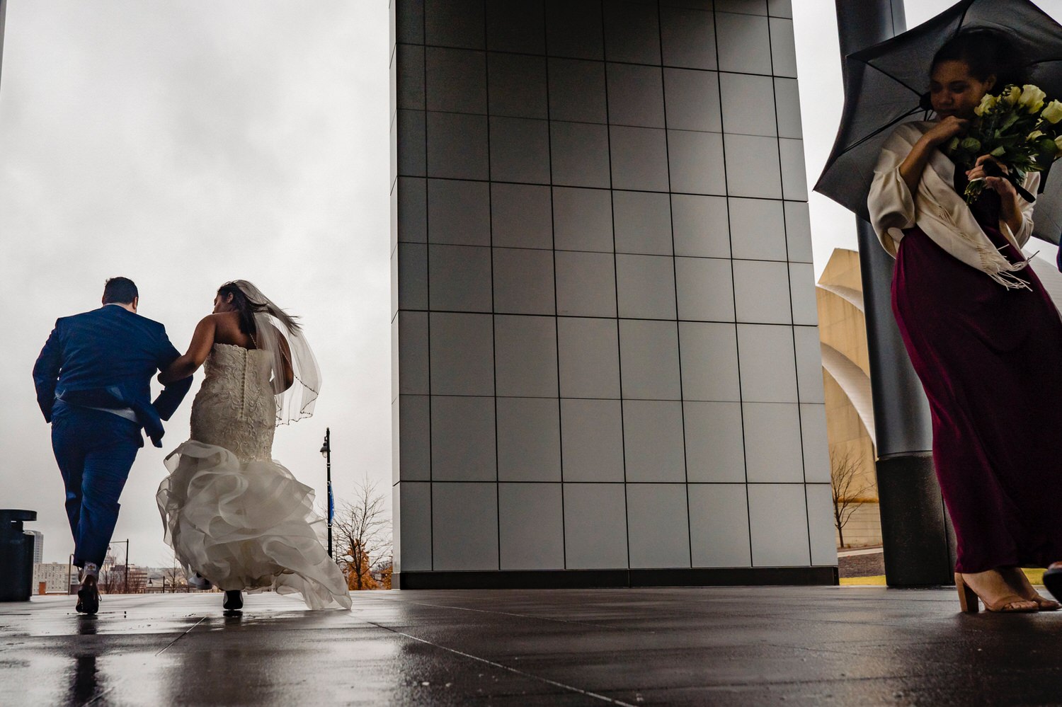 A colorful, candid picture of a bride and groom, holding hands walking off into the distance as a group of bridesmaids huddle under their umbrellas on a rainy winter wedding day in downtown Kansas City. 
