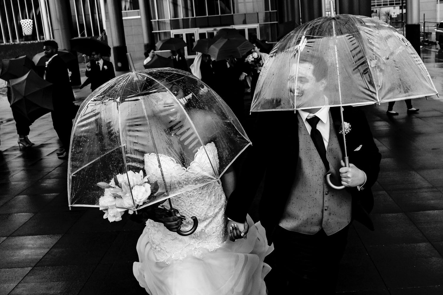 A candid black and white picture of a bride shrieking as a gust of wind tries to blow her umbrella away, her groom laughing underneath his own umbrella on their rainy winter wedding day in downtown Kansas City. 