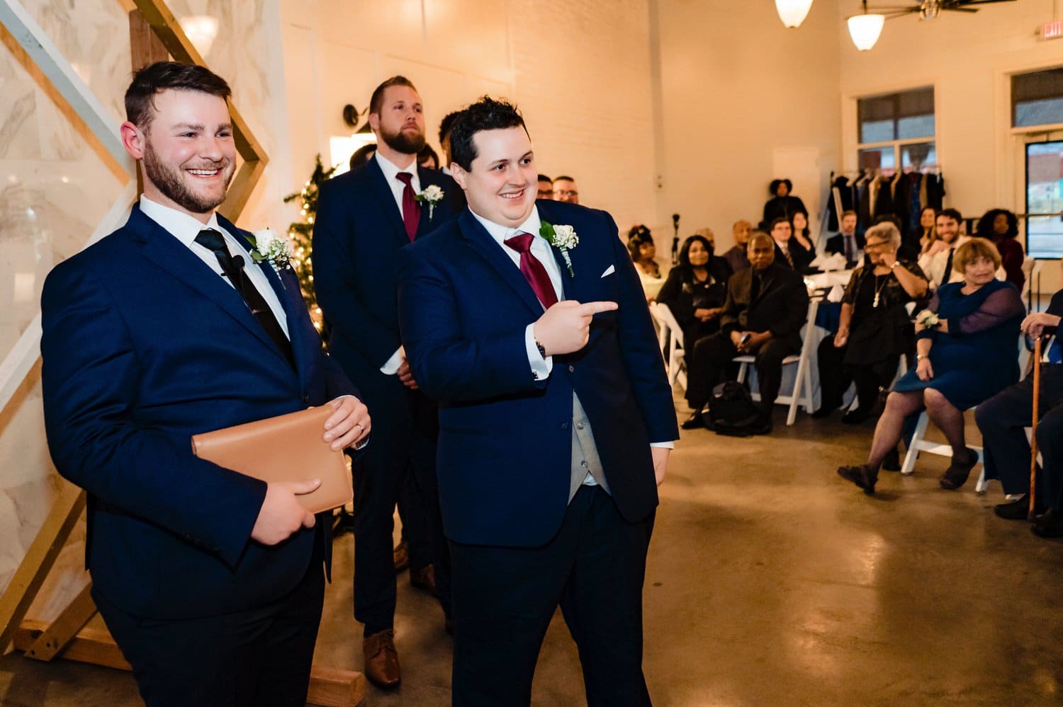 A colorful, candid picture of a groom smiling and pointing at his bride as she begins her walk down the aisle during their rainy winter wedding day at The Station in Kansas City. 