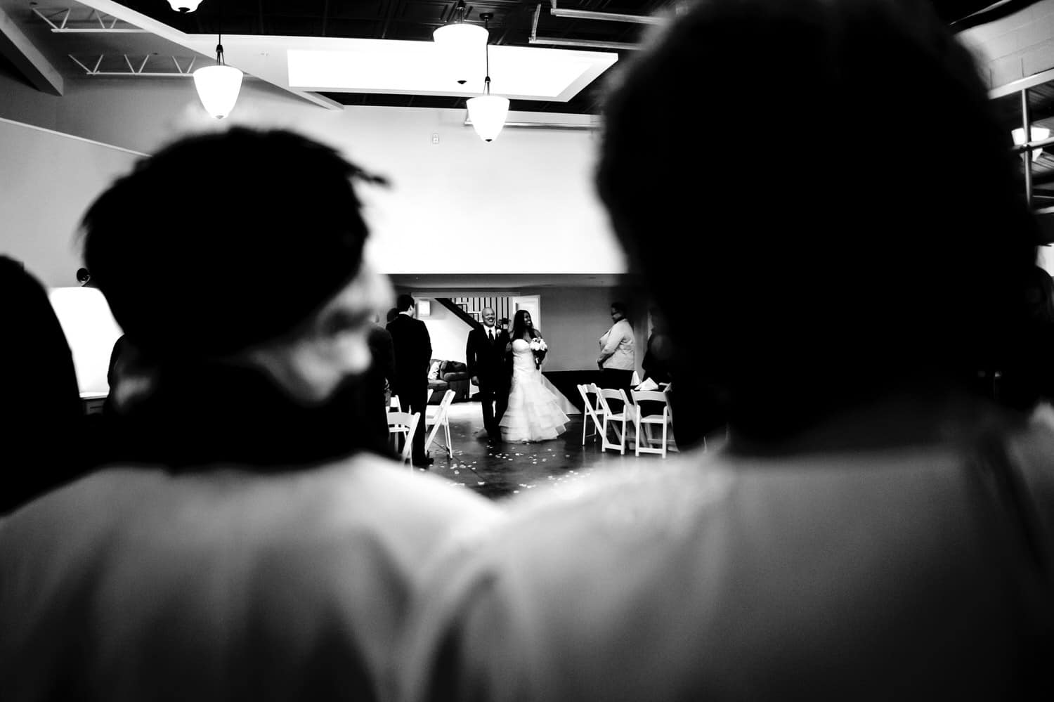 A candid black and white picture, taken over the shoulders of the bride's grandmas, of the bride and her dad beginning their walk down the aisle during her rainy winter wedding day at The Station in Kansas City. 