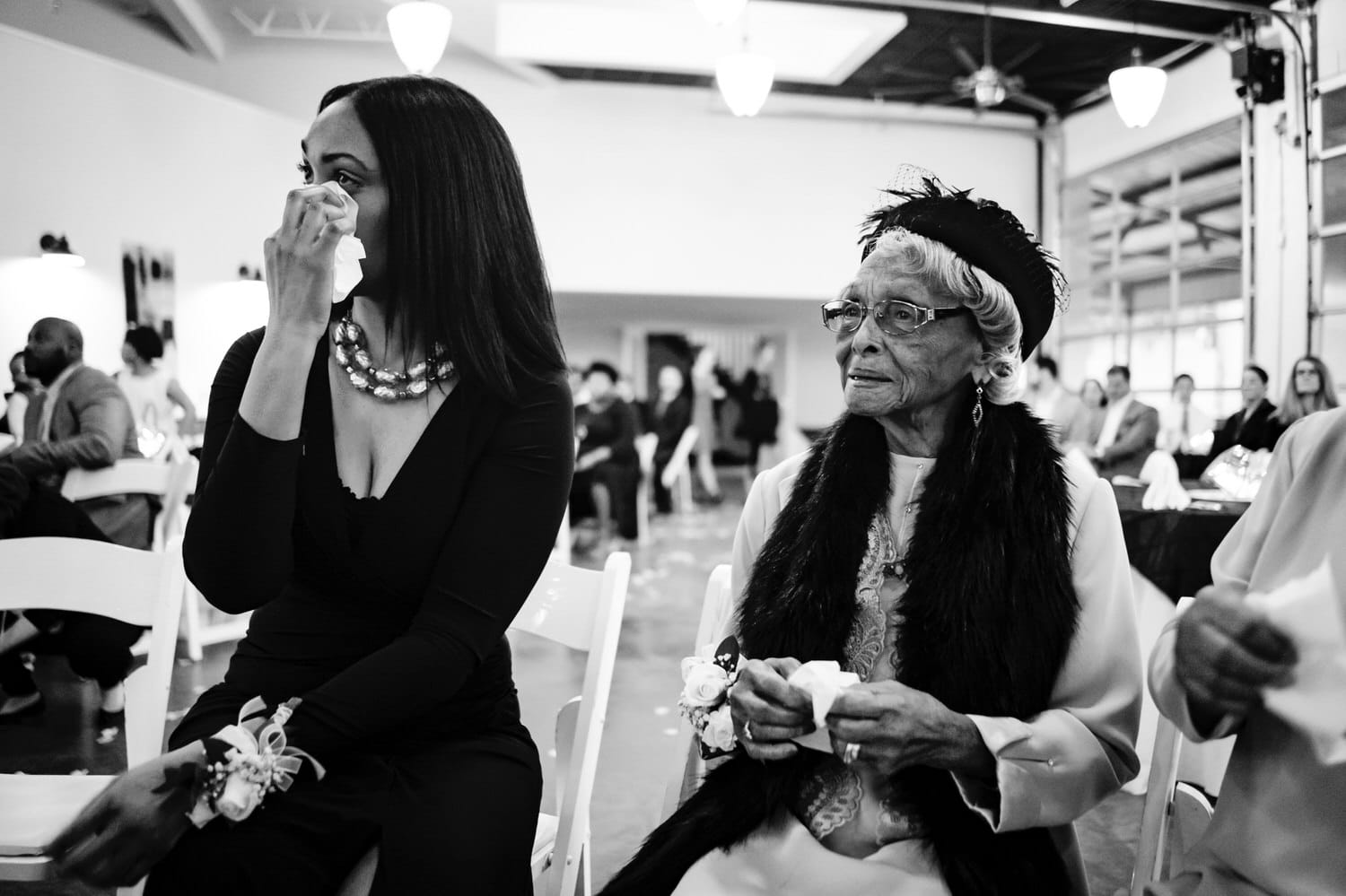 A close-up, candid, black and white picture of a bride's mom and grandma wiping tears from their eyes as a bride walks down the aisle during a winter wedding ceremony at The Station in Kansas City. 