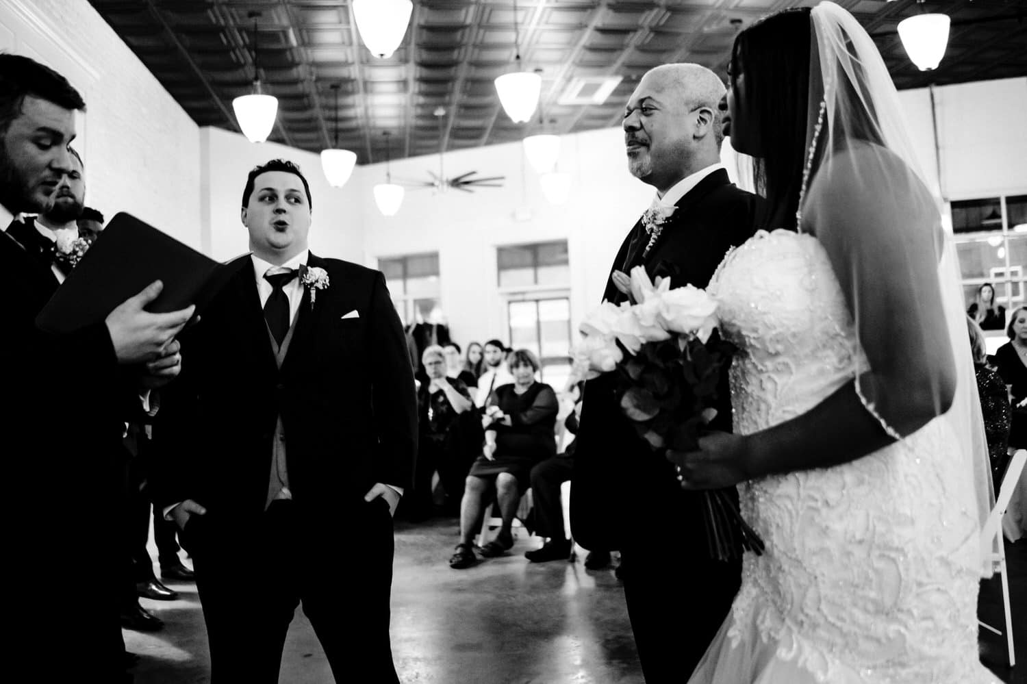 A candid black and white picture of a groom visibly exhaling as his bride and her father stand at the front of their wedding ceremony. 