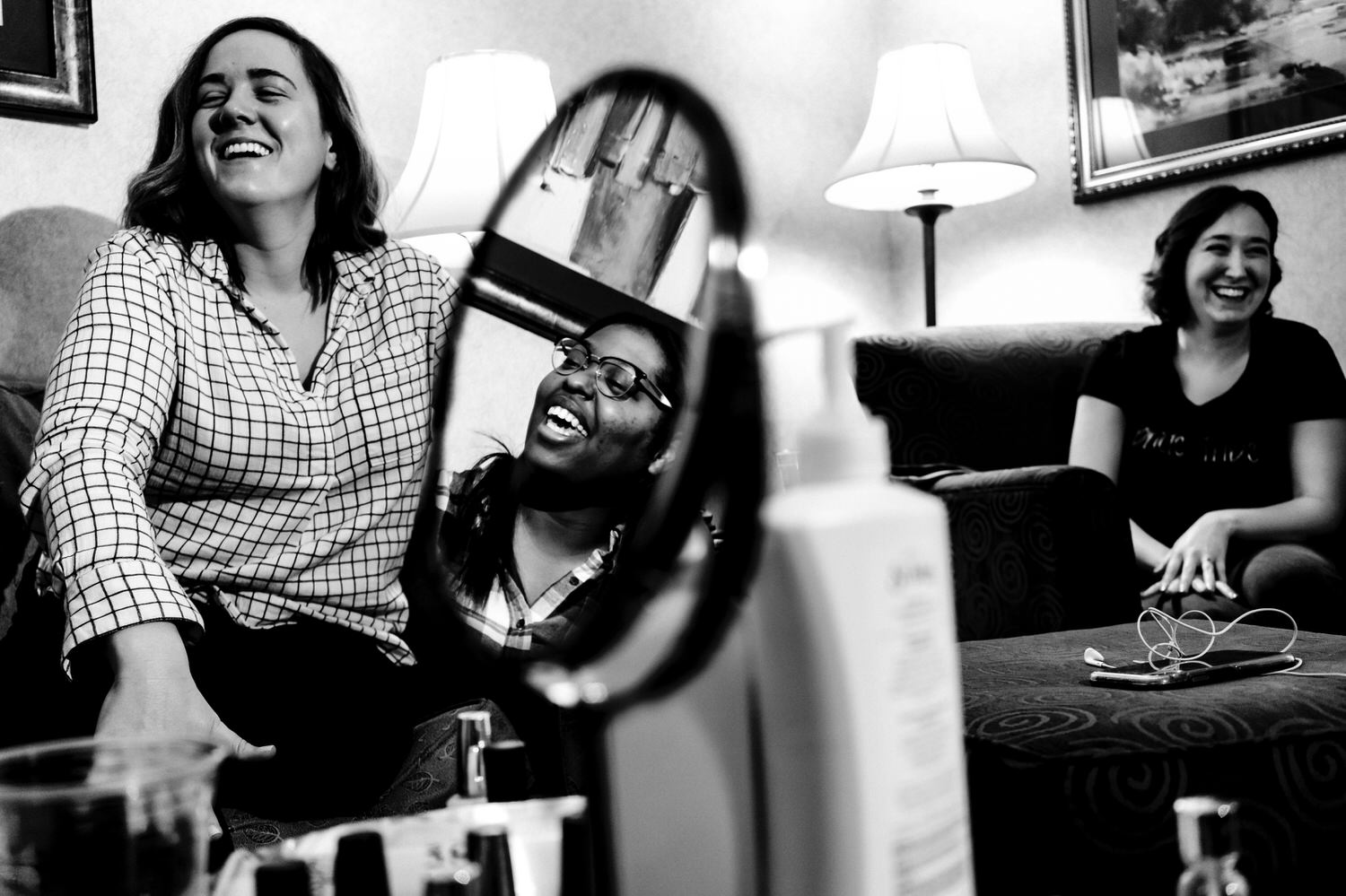 A candid black and white picture of two women, sitting on a couch laughing together as a mirror in the middle of a table shows the reflection of another woman laughing on a rainy winter wedding morning in Kansas City. 