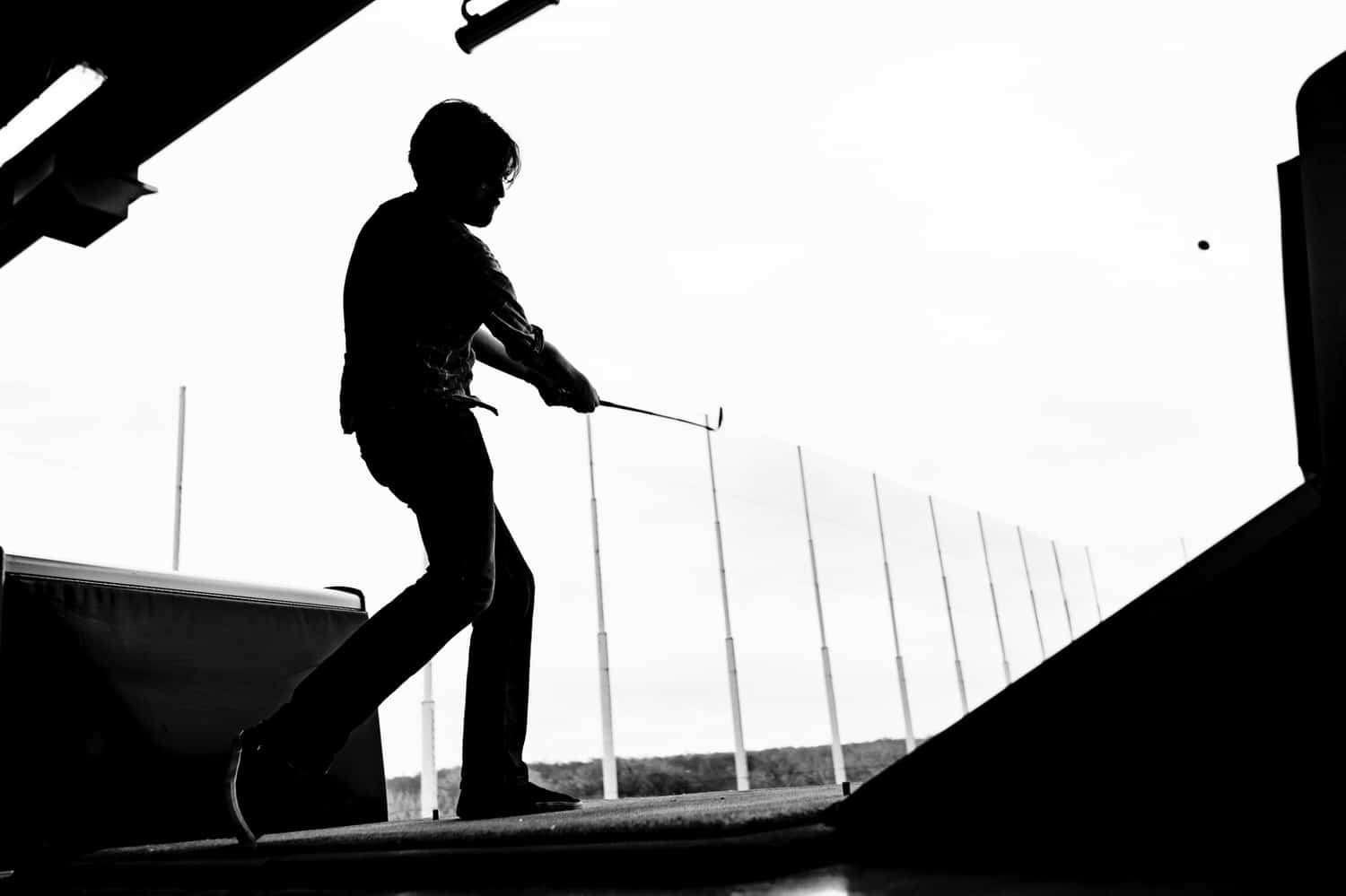 A black and white silhouetted picture taken behind a row of golf clubs of a man in a hat in the middle of his backswing at Top Golf on a rainy winter wedding morning in Kansas City. 
