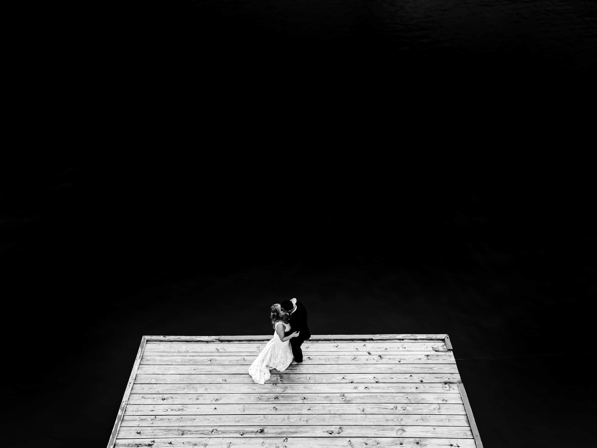 A dramatic black and white portrait of a bride and groom standing on a dock, sharing a kiss on their Kansas City Wedding Day.