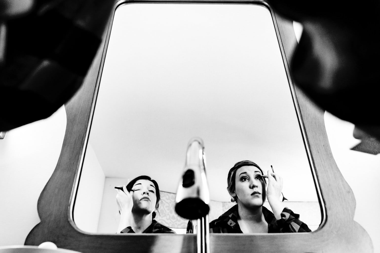 A candid black and white picture taken from directly behind and beneath two women applying eye shadow, visible in the mirror above a sink at The Elms. 