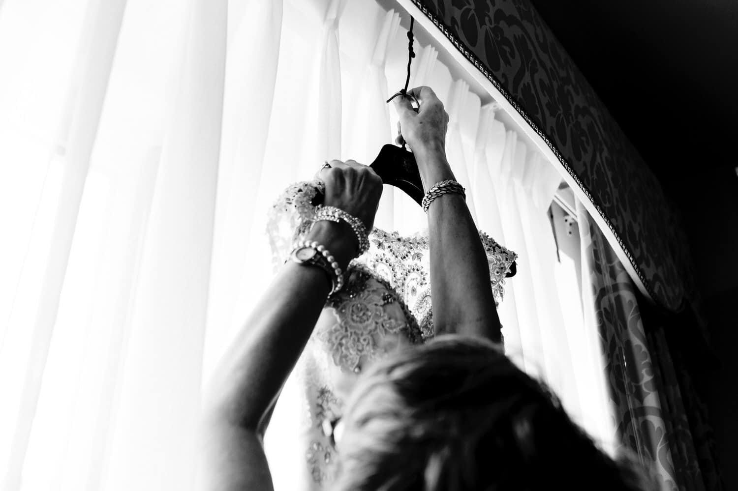 A candid black and white picture of a pair of women's hands reaching up to pull a wedding dress down from a window at The Elms. 