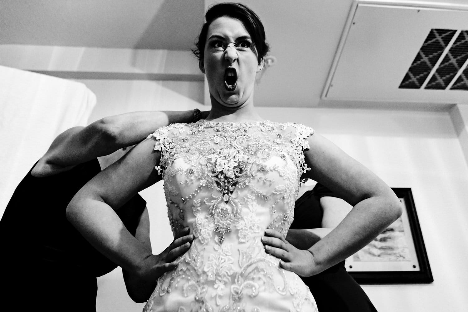A candid black and white picture of a bride, her hands on her hips, two women standing behind her zipping her up, screaming on her wedding day. 