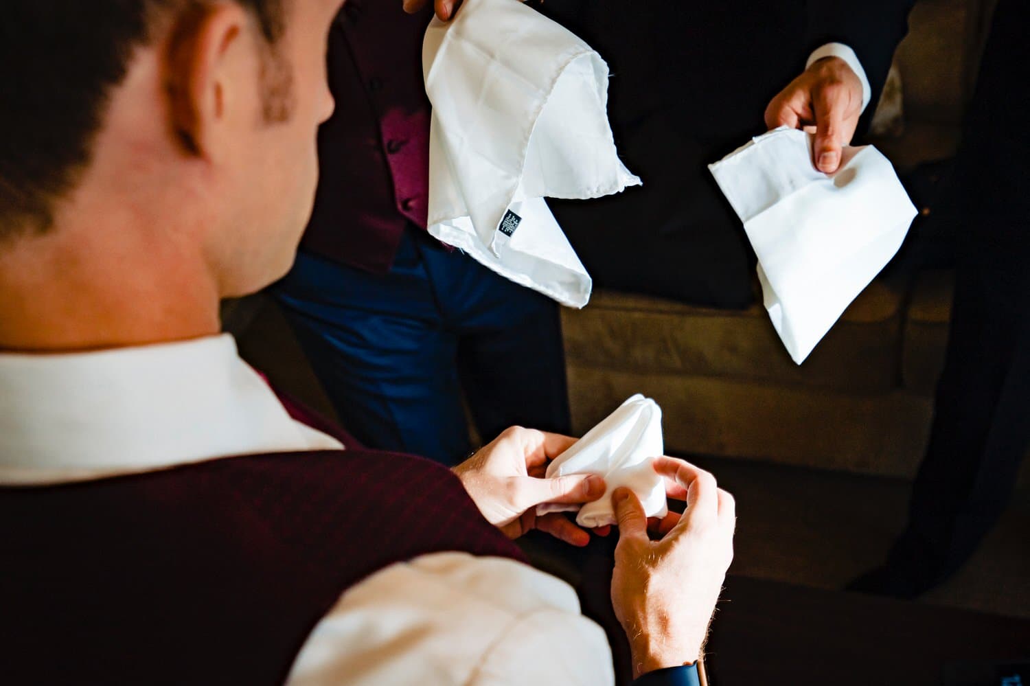 A colorful, candid picture of three men attempting to fold the pocket squares of their tuxedos on a wedding day. 