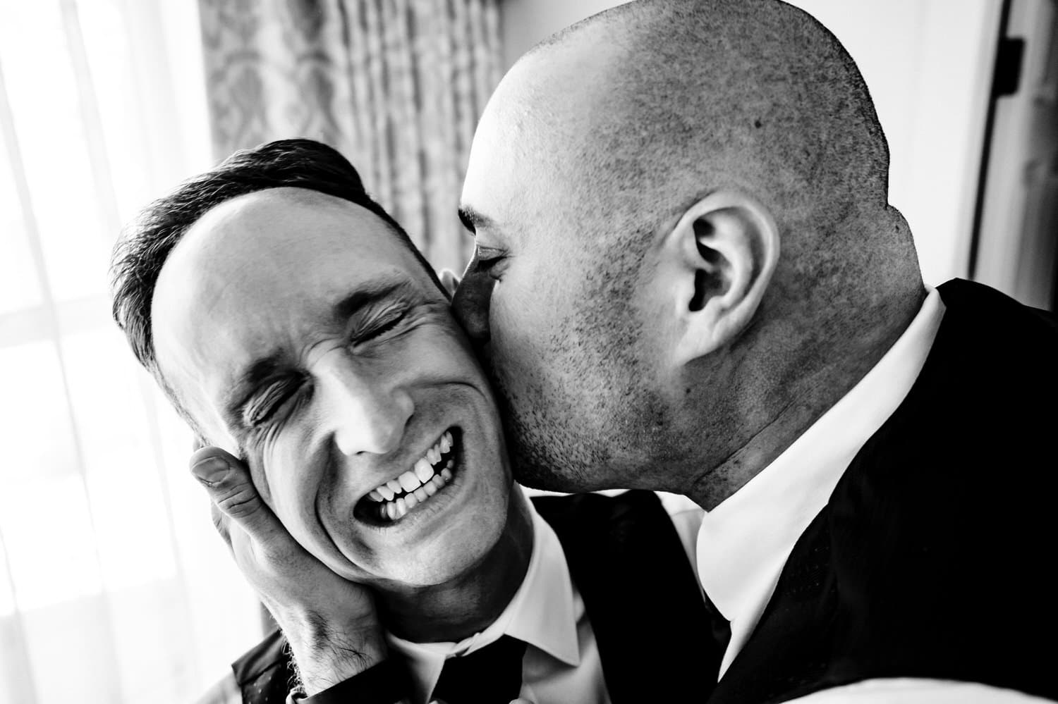 A candid black and white picture of a groomsman grabbing a groom's face and aggressively kissing him on the cheek as the groom laughs out loud. 