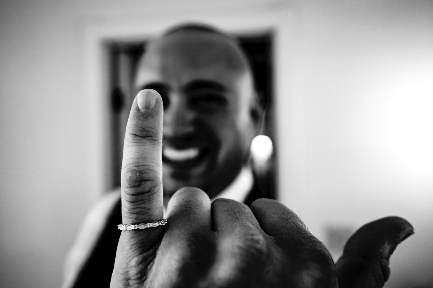 A black and white picture, focused on a dainty women's wedding band on a bulky man's pinky finger, the man smiling in the background. 