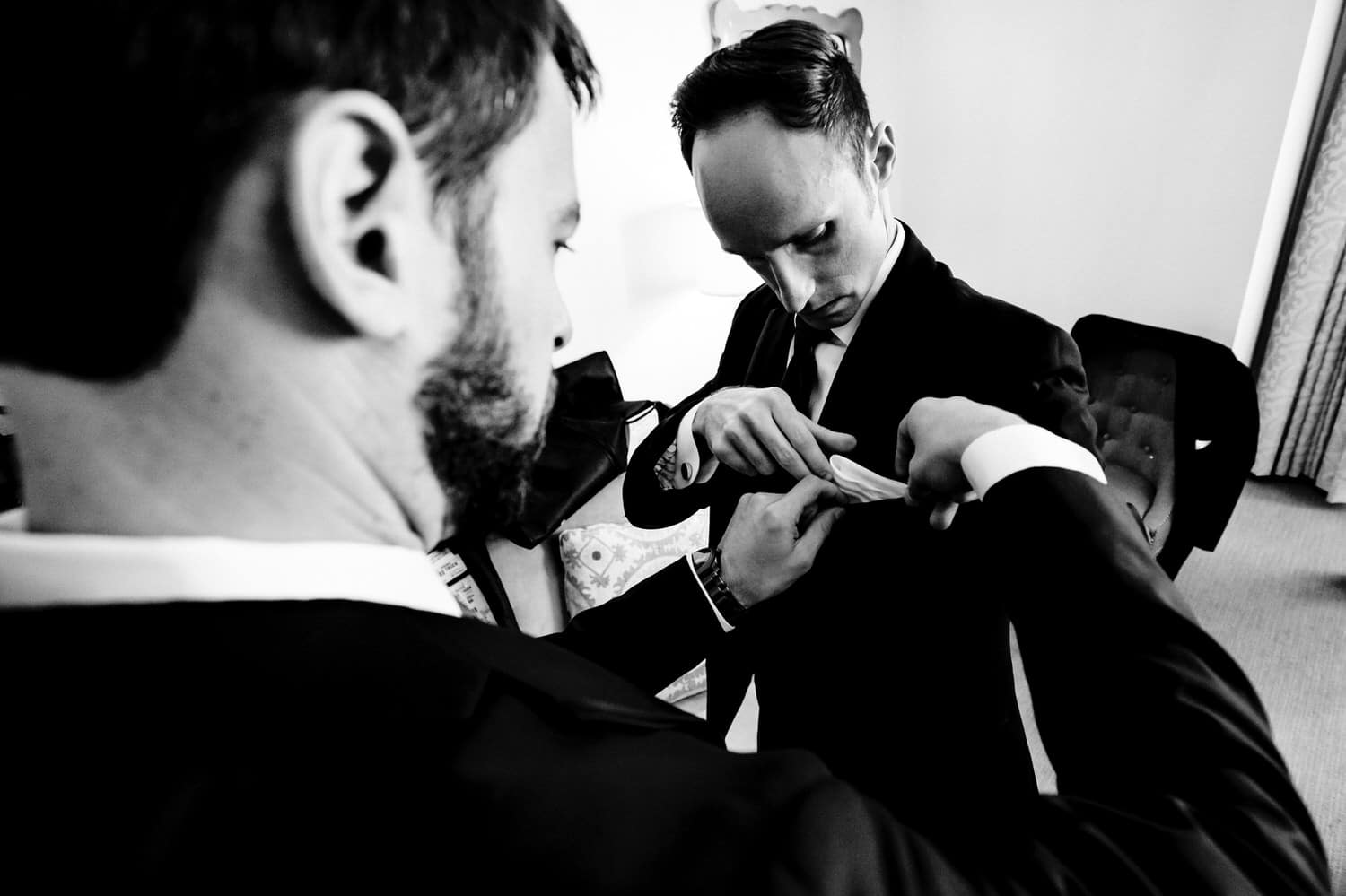 A candid black and white picture of a groom's best man helping him tuck the pocket square into his pocket. 