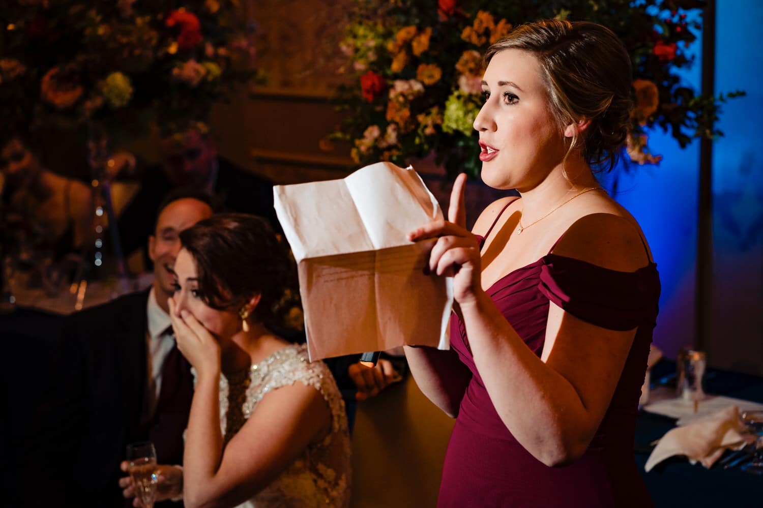 A colorful, candid picture, focused on a bridesmaid waving her finger around as she toasts the bride and groom, visible in the background with their hands over their mouths in surprise during a winter wedding reception at The Elms. 