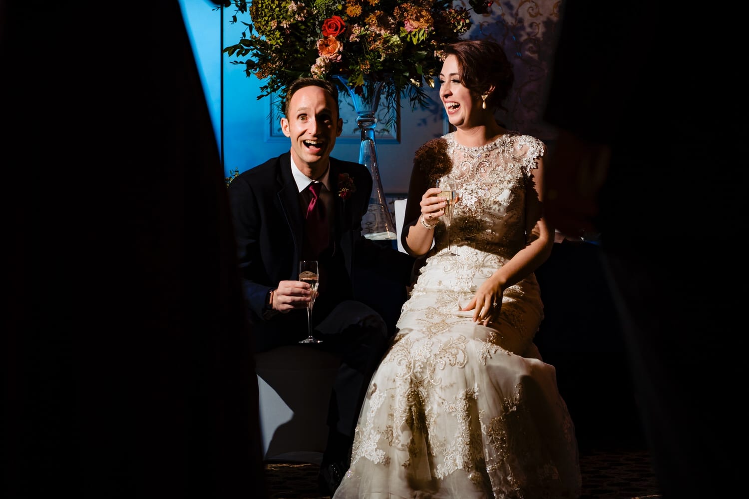 A colorful, candid picture of a bride and groom laughing hysterically as their wedding party toasts them during their wedding reception at The Elms. 